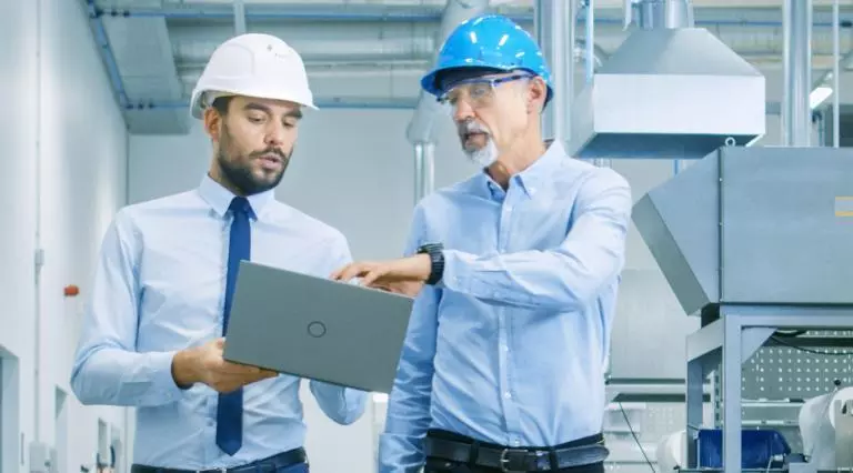 Two men walk through factory looking at laptop